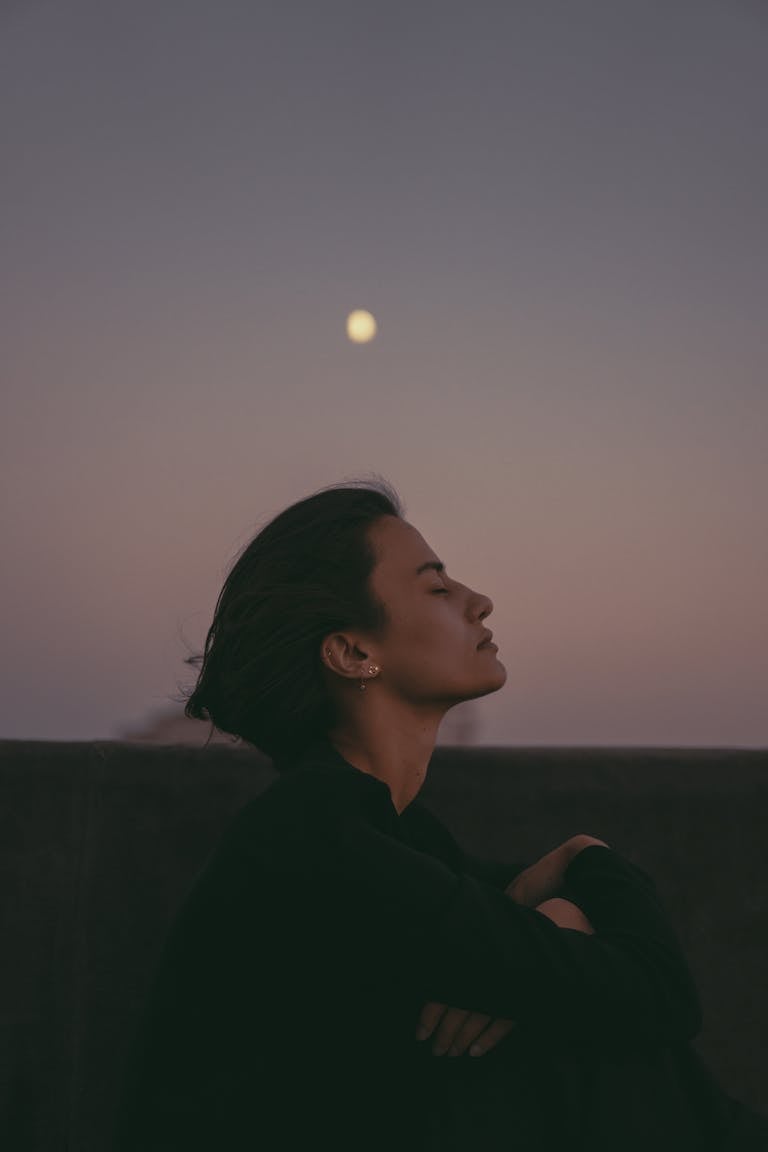 A serene woman enjoys a peaceful moment outdoors under the twilight sky with the moon visible.